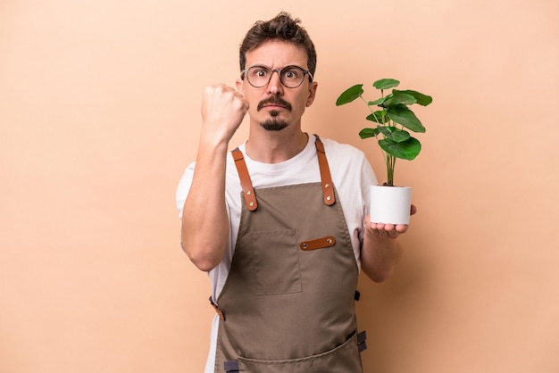 Jeune homme de jardinier caucasien tenant une plante isolée sur fond beige montrant le poing à la caméra expression faciale agressive