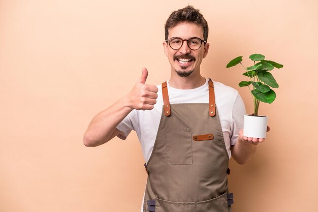 Jeune homme de jardinier caucasien tenant une plante isolée sur fond beige souriant et levant le pouce vers le haut