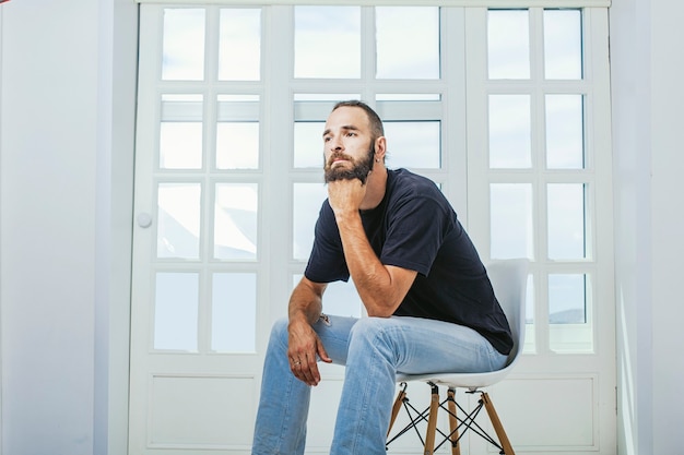 Jeune homme en jeans avec une barbe un hipster assis sur une chaise à l'arrière-plan