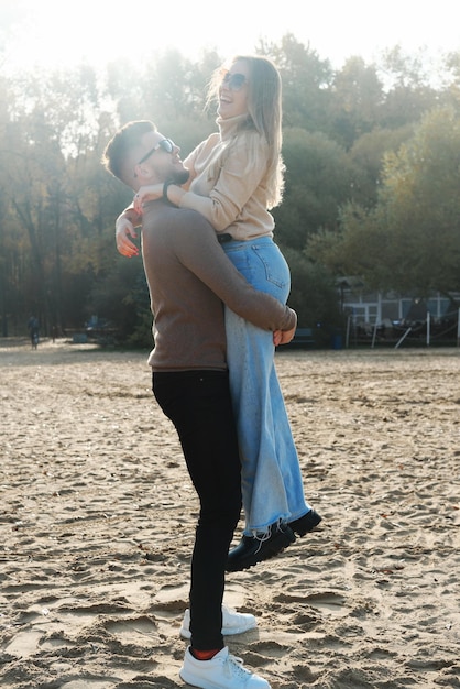 Un jeune homme en jeans foncé et un pull a pris une fille blonde dans ses bras Jeune couple s'amusant sur la plage