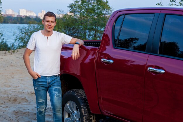 Photo jeune homme en jeans et t-shirt blanc près d'une voiture rouge sur le lac