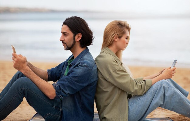 Photo un jeune homme et une jeune femme assis dos à dos et utilisant des smartphones à l'extérieur