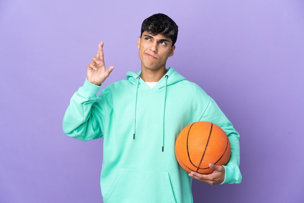 Jeune homme jouant au basket-ball sur mur violet isolé avec les doigts qui se croisent et souhaitant le meilleur
