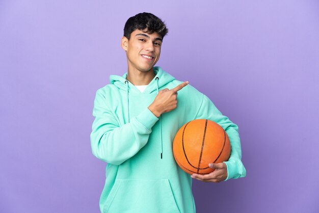 Jeune homme jouant au basket sur mur violet isolé pointant vers le côté pour présenter un produit
