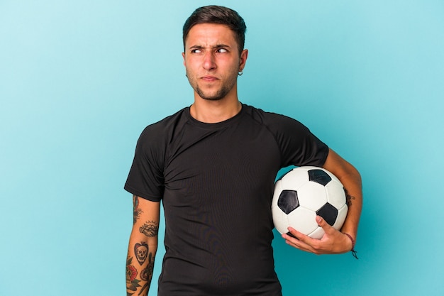 Jeune homme jouant au football tenant un ballon isolé sur fond bleu confus, se sent dubitatif et incertain.