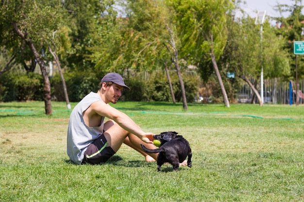 Jeune homme jouant avec un chien au parc Chiot noir mordant une balle de tennis avec le propriétaire assis sur l'herbe