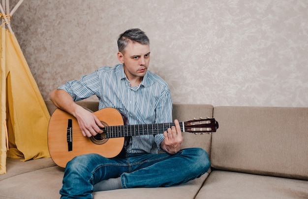 Un jeune homme joue de la guitare assis sur un canapé dans un salon lumineux. Un instrument de musique pour le concept de loisir ou de passe-temps. divertissements à domicile