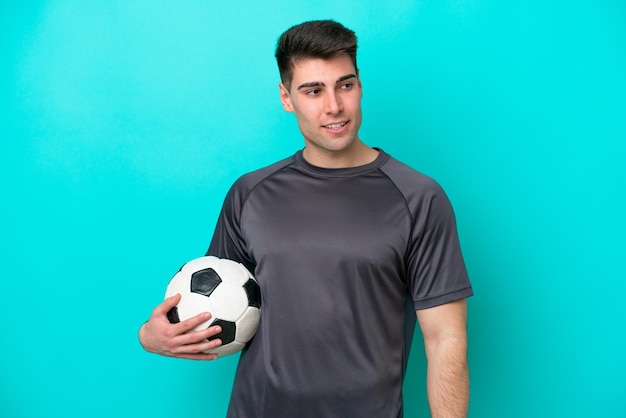 Jeune homme de joueur de football caucasien isolé sur fond bleu regardant sur le côté et souriant