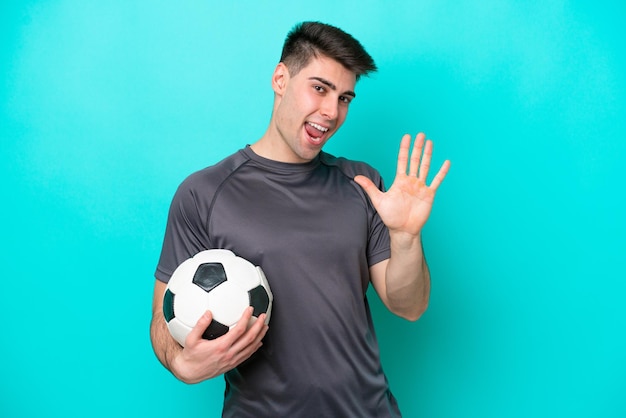 Jeune homme de joueur de football caucasien isolé sur fond bleu saluant avec la main avec une expression heureuse