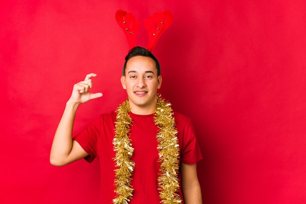 Jeune homme le jour de Noël tenant quelque chose de petit avec les index, souriant et confiant.