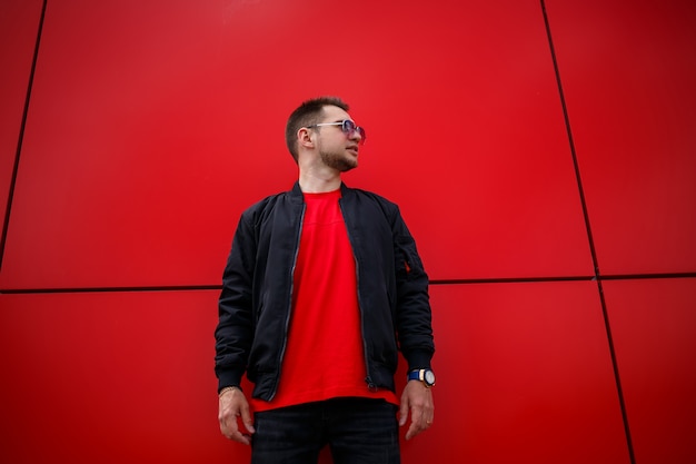 Jeune homme joyeux avec une barbe avec un sourire positif dans un t-shirt dans une veste noire à la mode dans un t-shirt posant près d'un bâtiment en métal rouge dans la ville. Heureux gars urbain dans la rue. Vêtements pour hommes