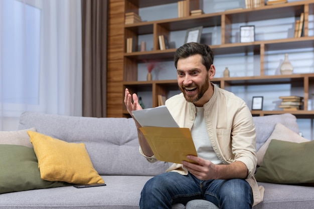 Un jeune homme joyeux est assis sur le canapé à la maison et tient une enveloppe avec une lettre dans ses mains