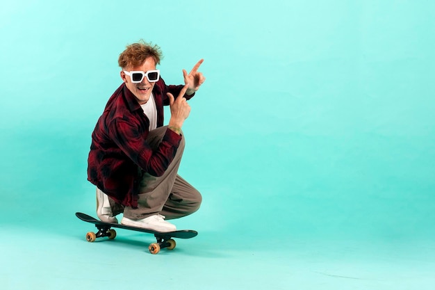 Un jeune homme joyeux fait du skateboard sur un fond bleu isolé, un patineur hipster avec des lunettes de soleil.