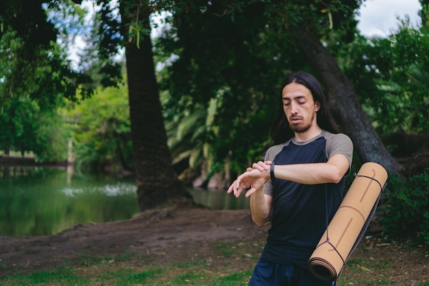 Jeune homme latin aux cheveux longs dans une forêt avec lac en regardant sa montre tout en portant un tapis de yoga sur son dos Espace de copie