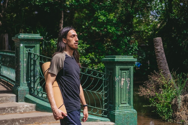 Jeune homme latin aux cheveux longs marchant sur un pont piétonnier avec un tapis de yoga sur le dos au bord d'une rivière Espace de copie