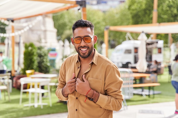 Jeune homme latin avec barbe portant une chemise orange et des lunettes de soleil portrait en plein air avec une terrasse de bar en arrière-plan