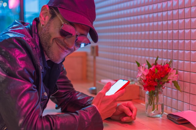 Jeune homme latin avec casquette noire, lunettes de soleil et veste en cuir regardant son smartphone dans un endroit avec des néons rouges