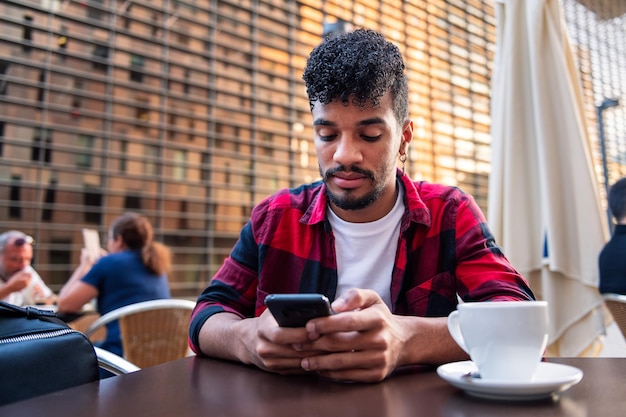 Jeune homme latin consultant son téléphone assis avec un café sur la terrasse d'un café, concept de technologie et mode de vie urbain