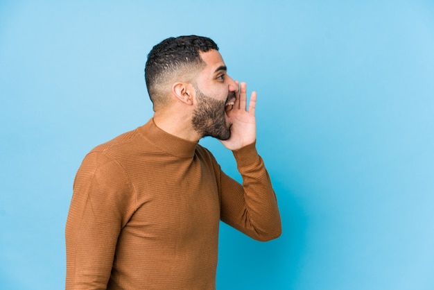 Jeune homme latin contre un mur bleu criant et tenant la paume près de la bouche ouverte.