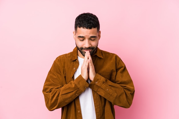 Jeune homme latin contre un mur rose isolé tenant par la main dans la prière près de la bouche, se sent confiant.