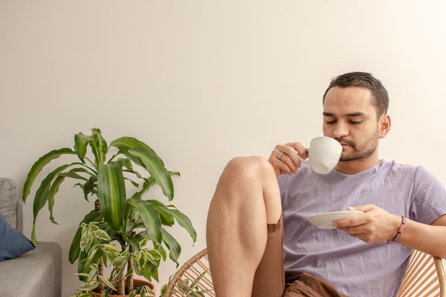 Jeune homme latin habillé avec désinvolture, se relaxant sur la chaise à la maison le matin tout en buvant une boisson chaude