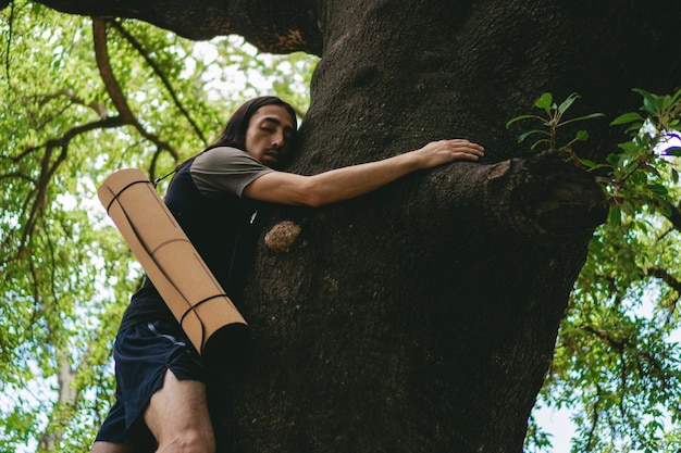 Jeune homme latin hippie aux cheveux longs et un tapis de yoga sur le dos serrant un tronc d'arbre comme thérapie