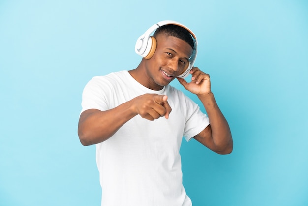 Jeune homme latin isolé sur un mur bleu à l'écoute de la musique et pointant vers l'avant