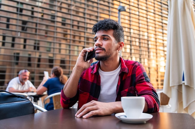 Jeune homme latin parlant au téléphone assis avec un café sur la terrasse d'un café, concept de technologie et mode de vie urbain