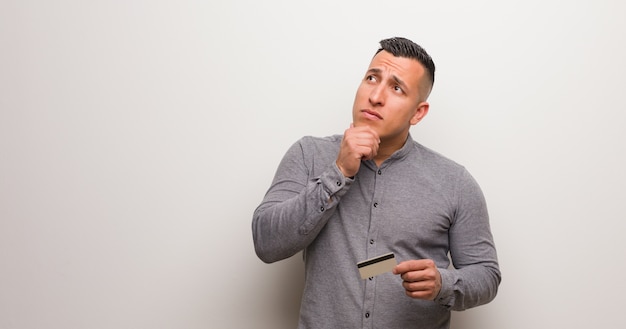 Jeune homme latin tenant une carte de crédit doutant et confus