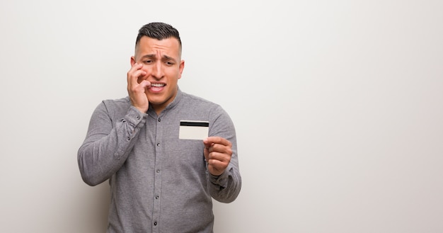 Jeune homme latin tenant une carte de crédit se ronger les ongles, nerveux et très inquiet