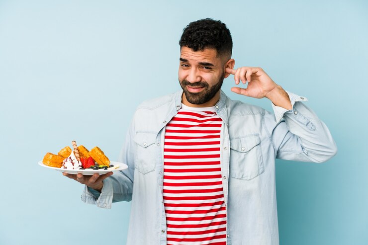 Photo jeune homme latin tenant une gaufre isolé couvrant les oreilles avec les mains.