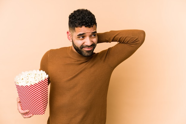 Jeune homme latin tenant un pop corn isolé toucher l'arrière de la tête, penser et faire un choix.