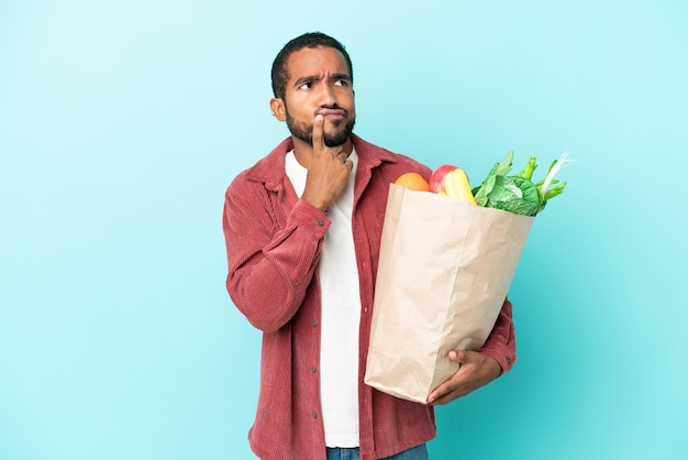 Jeune homme latin tenant un sac d'épicerie isolé sur fond bleu ayant des doutes en levant les yeux