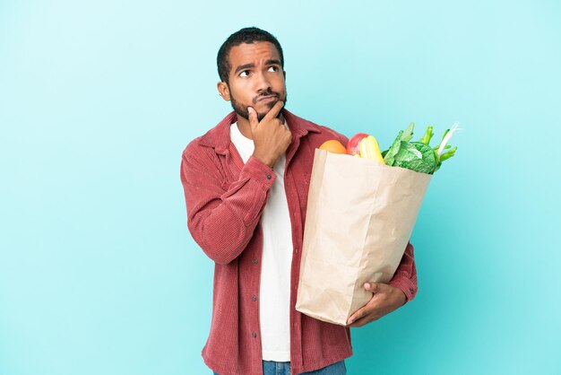 Jeune homme latin tenant un sac d'épicerie isolé sur fond bleu ayant des doutes