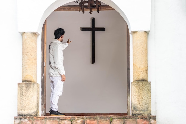 Jeune homme latin vêtu de blanc debout à la porte d'une église chrétienne regardant la croix