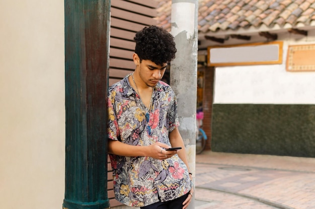 Un jeune homme latino-américain aux cheveux bouclés vérifie son téléphone portable dans la rue en s'appuyant sur un vieux poteau de lumière de rue