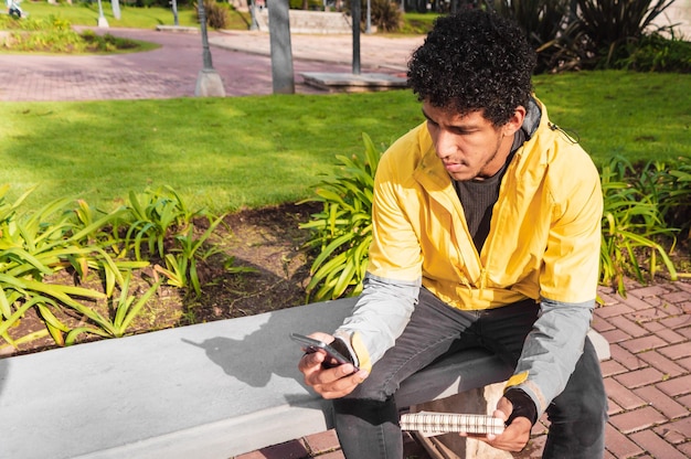 Jeune homme latino assis sur une place urbaine avec un téléphone et un ordinateur portable dans ses mains vêtu de jaune concept de technologie