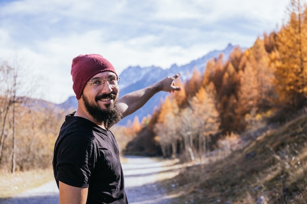 Jeune homme avec des lunettes et une barbe vêtu d'un Tshirt noir et un chapeau rouge pointant vers les montagnes