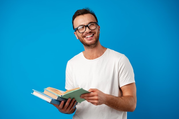 Jeune homme avec des lunettes debout et lisant un livre sur fond bleu