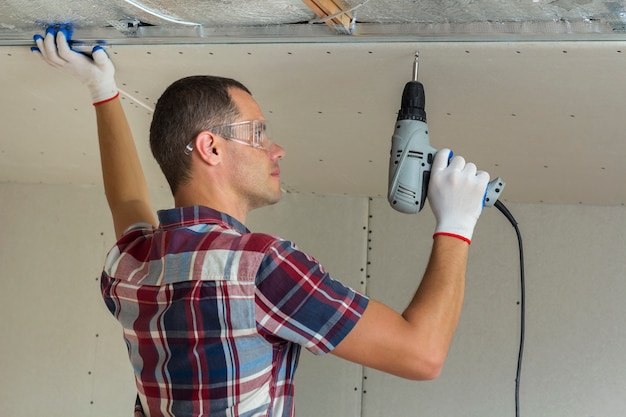 Jeune homme à lunettes fixant le plafond suspendu pour cloisons sèches au cadre métallique à l'aide d'un tournevis électrique pour fixation au plafond.