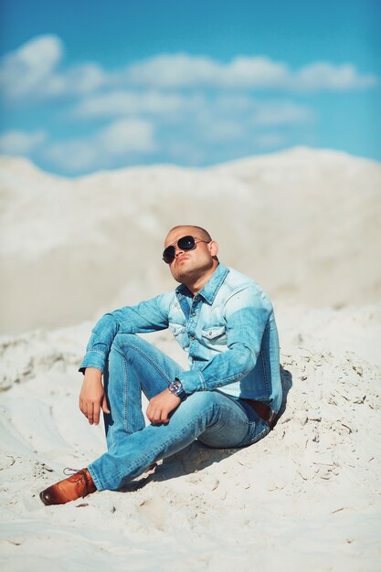 Jeune homme à lunettes se trouve dans des vêtements jeans sur le sable à Dubaï