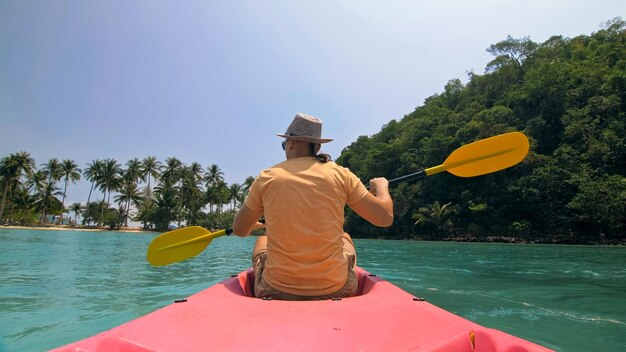 Photo jeune homme avec des lunettes de soleil et un chapeau rangées canoë en plastique rose le long de la mer contre les îles vallonnées vertes
