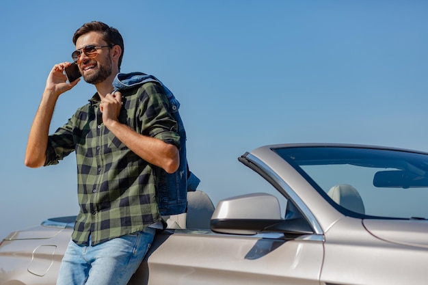 Jeune homme à lunettes de soleil debout à la voiture et ayant une conversation au téléphone