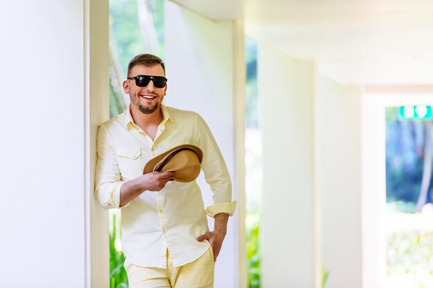 Jeune homme à lunettes de soleil portant un chapeau jaune et des vêtements décontractés s'amuser en journée ensoleillée. Fond tropical.