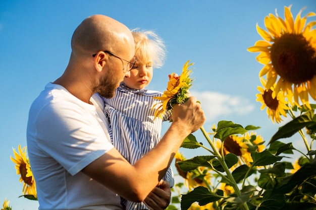 Jeune homme à lunettes tient un enfant dans ses bras et regarde les fleurs d'un tournesol sur un champ de tournesol