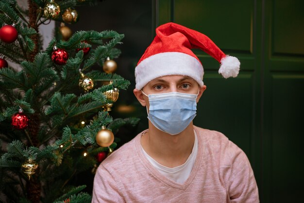 Jeune homme à la maison près d'un arbre de Noël portant un masque et un chapeau de père Noël regarde la caméra