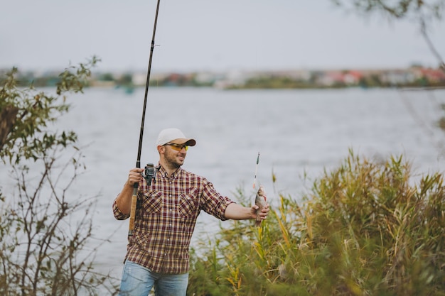 Un jeune homme mal rasé en chemise à carreaux, casquette et lunettes de soleil a sorti une canne à pêche avec du poisson pêché et se réjouit au bord du lac près des arbustes et des roseaux. Mode de vie, loisirs, concept de loisirs de pêcheur