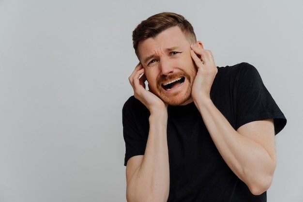 Jeune homme mal rasé effrayé en t-shirt noir ayant peur de quelque chose et criant, regardant avec une expression effrayée quelque chose en se tenant debout sur fond gris. Concept de peur et d'anxiété