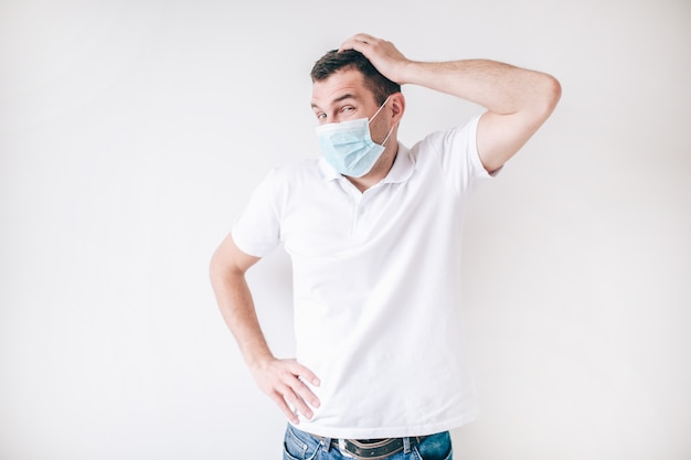 Jeune homme malade isolé sur mur blanc. Guy porte un masque facial de protection médicale pour éviter la grippe et les virus. Un homme confus tient la main sur la tête et regarde vers la gauche.