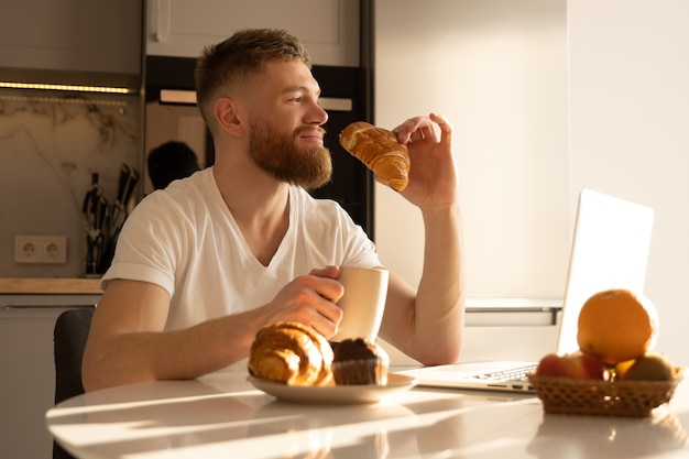 Jeune homme mangeant un croissant et buvant du thé ou du café au petit-déjeuner. Homme barbu européen souriant assis à table avec de la nourriture et un ordinateur portable. Intérieur de cuisine dans un appartement moderne. Temps du matin ensoleillé